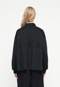 Sweatshirt preto com costas plissadas, colarinho canelado e mangas longas. Apresenta um material superior liso com plissados texturizados na parte inferior.
