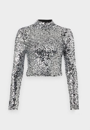 Kuttet langermet topp laget av sølvpaljetter med høy hals. Stoffet er blankt med en teksturert overflate som reflekterer lys.