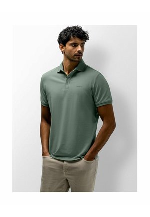 Man with curly hair wearing green short-sleeve polo shirt and beige pants, standing with hands in pockets, looking to the side against white background.