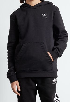 Dziecko ubrane w czarną bluzę z kapturem Adidas oraz dopasowane czarne spodnie z białymi logo i paskami Adidas, jedna ręka w kieszeni bluzy.