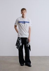 Bela majica s modrimi črtami in logotipom, črne razširjene hlače ter siva karo srajca, vezana okoli pasu. Črni čevlji. Minimalistična podlaga.