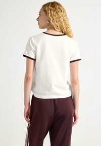 Kortærmet hvid t-shirt med mørkebrune kanter, lavet af bomuldsstof, afslappet pasform, synlige sømdetaljer, parret med bordeauxfarvede bukser.