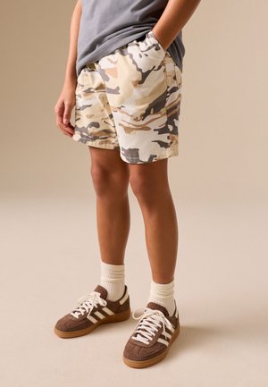 Barn iført beige og brun camouflage shorts, grå t-shirt, hvide sokker og brune sneakers står med hænderne i lommerne.
