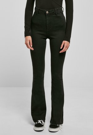 Personne portant un jean noir taille haute évasé, un haut noir à manches longues et des baskets noires et blanches sur un fond uni.