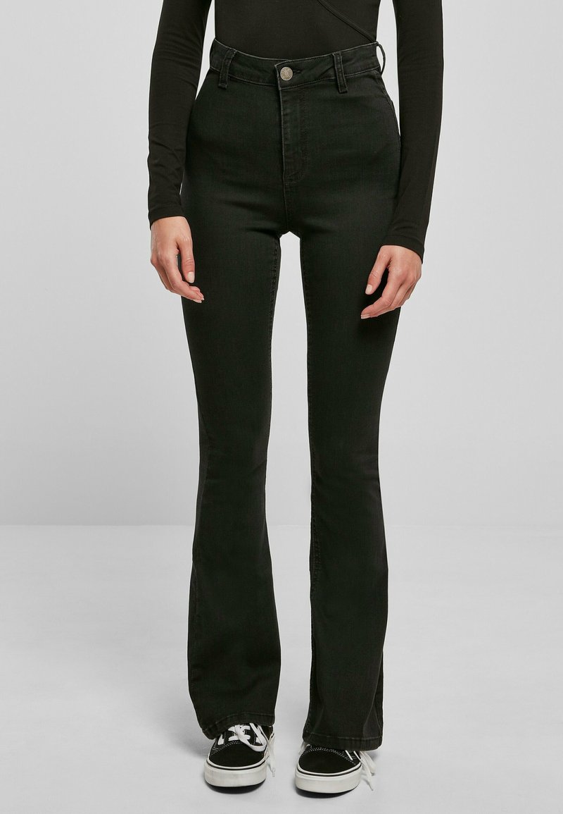 Personne portant un jean noir taille haute évasé, un haut noir à manches longues et des baskets noires et blanches sur un fond uni.