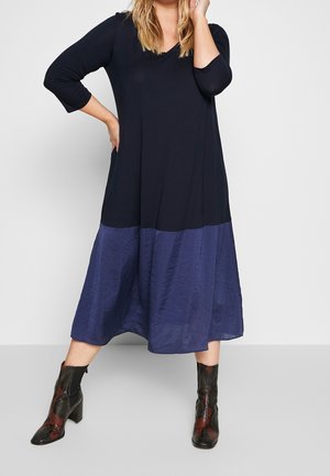 Femme portant une longue robe noire et bleu foncé avec des manches trois-quarts et des bottines noires à talons hauts avec des motifs rouges.