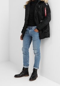 Veste matelassée noire avec capuche en fausse fourrure, poches zippées, associée à un jean slim bleu clair et des bottes noires à lacets.
