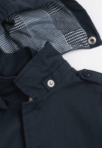 Veste bleu marine avec un col haut, des fermetures à boutons et à pression à l'avant, et une doublure intérieure à motif dans des tons de bleu et de blanc.