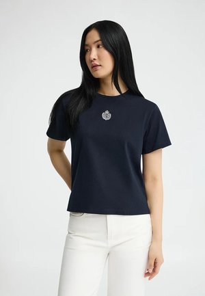 Kobieta z długimi czarnymi włosami ubrana w granatową koszulkę z krótkim rękawem z małym emblematem, w białych spodniach, stojąca na jednolitym tle.