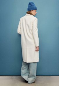 Cappotto bianco in lana dal taglio dritto, abbinato a jeans larghi azzurri e a un berretto a maglia blu, su uno sfondo blu solido.