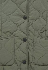 Tissu matelassé vert olive avec boutons noirs et deux poches avant, doté d’un motif de couture ondulé.