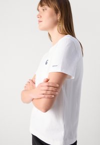 Jauna moteris su šviesiai rudais plaukais, vilkinti baltą GANT marškinėlį, rankos sulenktos kryžmiškai, žiūri į priekį priešais baltą foną.