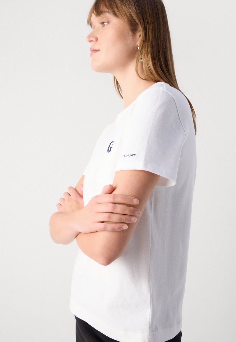 Junge Frau mit hellbraunen Haaren, die ein weißes GANT-T-Shirt trägt, mit verschränkten Armen und schaut auf ein weißes Hintergrundbild.