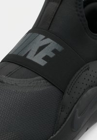 Zwarte sneaker met een gestructureerde bovenkant, elastische band met "NIKE" branding en geperforeerde accenten aan de zijkant.