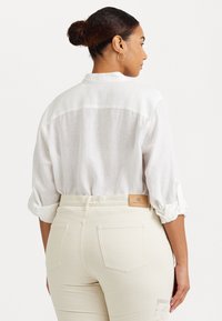 Camisa de lino blanca con mangas remangadas, que muestra un ajuste relajado, combinada con jeans crema que presentan un parche de piel marrón en la parte trasera.