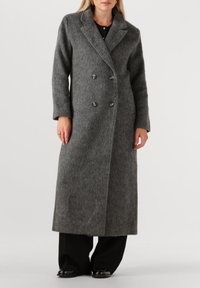 Grijze wollen jas met een dubbelzijdig ontwerp, notch-kraag en vier knopen. Tot onder de knie, gecombineerd met brede zwarte broeken en schoenen.