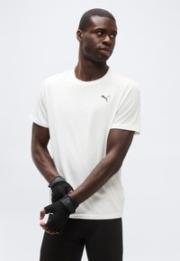 Άνδρας που φοράει λευκό T-shirt Puma και μαύρα γάντια άσκησης χωρίς δάχτυλα, κοιτώντας προς τα αριστερά του με φόντο έναν απλό ανοιχτόχρωμο τοίχο.