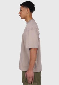 Helles, braunes Baumwoll-T-Shirt mit kurzen Ärmeln, entspannter Passform, Rundhalsausschnitt und weißem Grafikdruck auf der Vorderseite. Getragen mit olivgrünen Hosen.