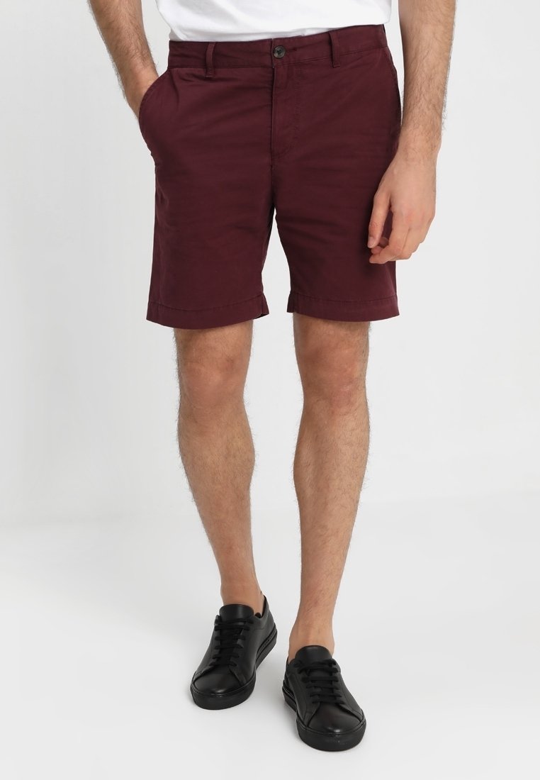 Homme portant un short rouge foncé avec des poches avant et des baskets noires basses, debout devant un fond clair uni.