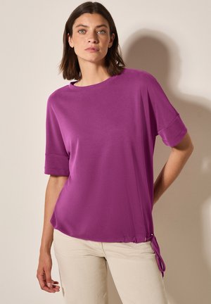 Haut violet à manches courtes avec un col rond et un détail à cordon sur le côté. Tissu doux avec une texture lisse. Associé à un pantalon beige clair.