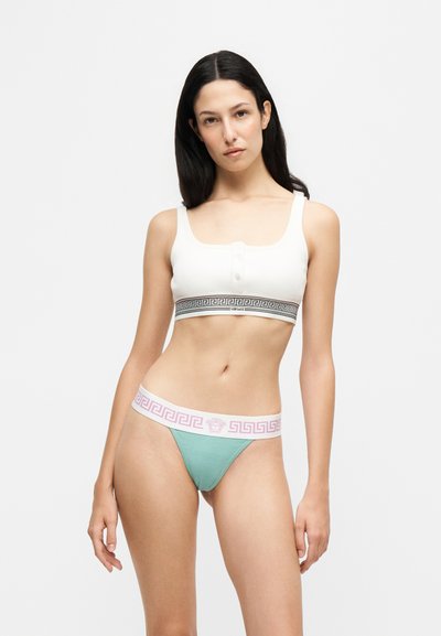Femme aux longs cheveux foncés portant un crop top blanc et un sous-vêtement vert avec des bandes élastiques brandées, debout devant un fond clair uni.