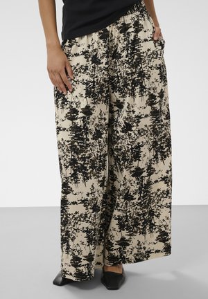 Pantalon beige à jambes larges avec motif arbre abstrait noir, porté avec un haut noir et des chaussures noires pointues, mains dans les poches.