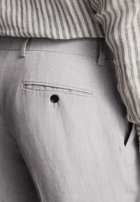 Gros plan sur un pantalon gris clair sur mesure avec une chemise rayée rentrée, montrant une poche arrière avec un bouton et des passants de ceinture.