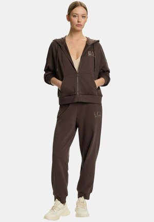 Donna in piedi con le mani nelle tasche, indossa una felpa con zip marrone scuro e pantaloni jogger abbinati con sneakers beige su sfondo bianco.