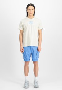 Lys beige bomulds t-shirt med et blåt grafisk logo, parret med blå cargo-shorts med sidelommer og grå mønstrede sneakers.