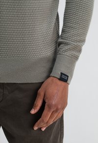 Lichtgrijze gebreide trui met een textuurpatroon, ribgebreide boorden en zoom. Opvallend logootasje op de mouw. Donkerbruine broek is zichtbaar.