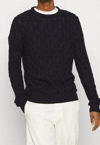 Marinblå stickad tröja med diamantmönster, rundad halsringning och ribbade muddar och nederkant, bärs över en vit skjorta. Slät textur.