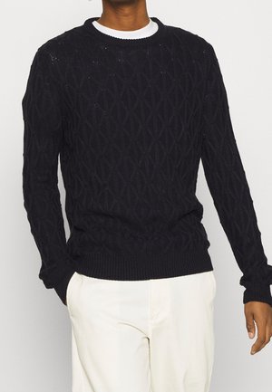 Pull en tricot bleu marine avec un motif en diamant, col rond et bords côtelés, porté par-dessus une chemise blanche. Texture lisse.