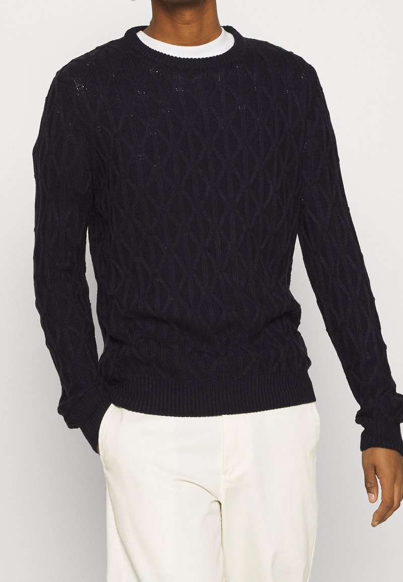 Marinblå stickad tröja med diamantmönster, rundad halsringning och ribbade muddar och nederkant, bärs över en vit skjorta. Slät textur.