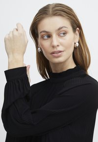 Haut noir plissé à col montant avec de longues manches bouffantes. Modèle portant des boucles d'oreilles rondes en argent, montrant un teint de peau lisse.