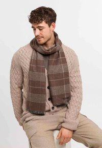 Brunternet tørklæde med frynsede kanter, lavet af et blødt materiale, båret over en beige strikket sweater med strukturerede mønstre og knapper.
