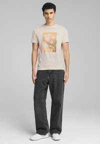T-shirt beige chiaro con stampa grafica raffigurante un leone e la parola "PUMA", abbinato a jeans larghi scuri e sneakers bianche.