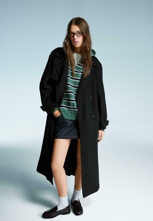 Sort dobbeltbrystet frakke over en grøn og hvid stribet hoodie, denimskørt, grå sokker og mørke loafers. Enkel baggrund.