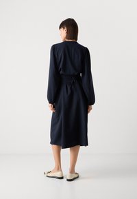 Robe rayée bleu marine avec de longues manches bouffantes, taille élastique et lien dans le dos. Associée à des ballerines crème et une texture lisse.