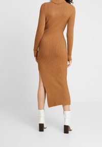 Robe en maille côtelée marron, avec des manches longues et une fente sur le côté, portée avec des bottes à talons blanches. Tissu en tricot texturé.