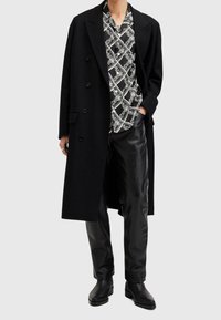 Cappotto di lana nero, camicia a righe nere e bianche, pantaloni di pelle neri e stivali neri. La camicia presenta un design geometrico.