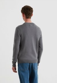 Mężczyzna tyłem w szarym swetrze z dzianiny i niebieskich dżinsach na tle jasnego, jednolitego tła.