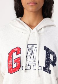Haut à capuche gris clair avec un grand logo "GAP", ornée de motifs cachemire rouges, blancs et bleu marine. Comprend une capuche avec cordon et un tissu texturé.
