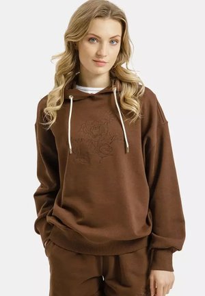 Kobieta o blond włosach, ubrana w brązową bluzę z kapturem i dopasowane spodnie, z delikatnym kwiatowym haftem oraz białymi sznurkami przy kapturze.