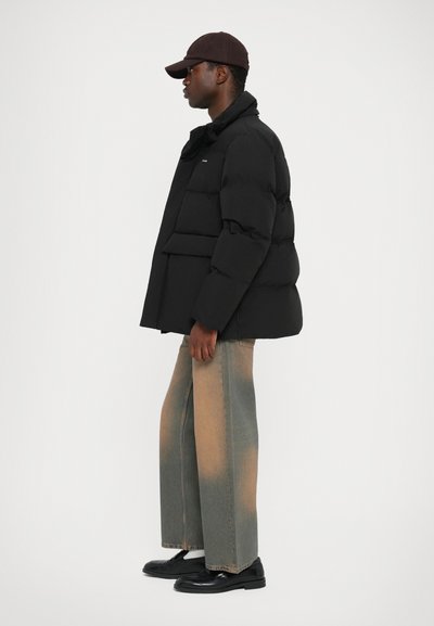 Black puffer jacket with a high collar and horizontal stitching, paired with wide-leg faded denim jeans and black shoes. Brown cap.