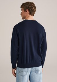 Pull bleu marine à col rond, manches longues, avec poignets et ourlet côtelés. Porté avec un jean bleu clair. Texture lisse.