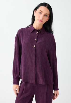 Femme aux cheveux noirs et lisses portant une chemise texturée manches longues violet foncé avec des boutons dorés et un pantalon assorti sur fond blanc.
