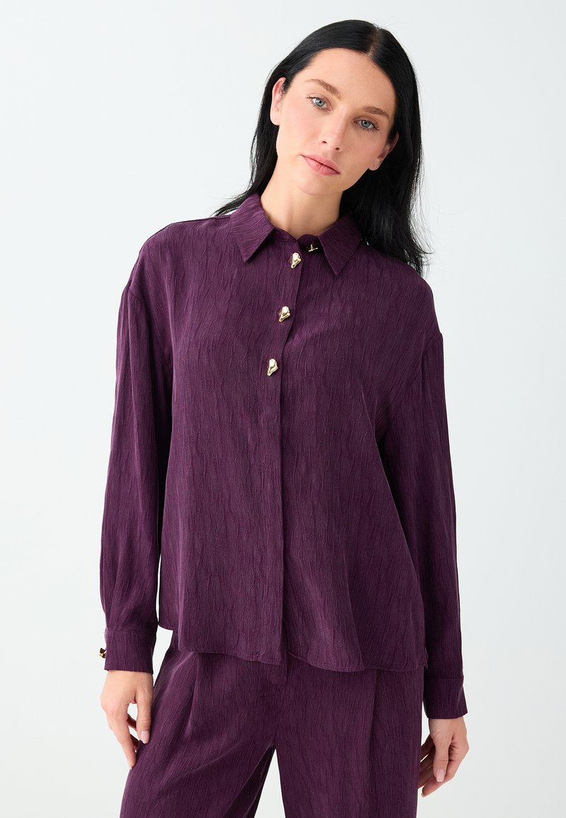 Femme aux cheveux noirs et lisses portant une chemise texturée manches longues violet foncé avec des boutons dorés et un pantalon assorti sur fond blanc.