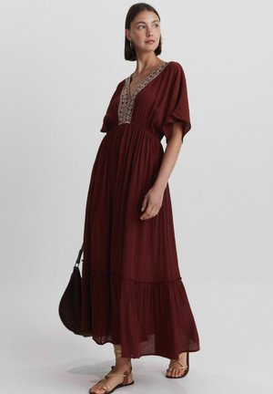 Femme portant une longue robe bordeaux avec une broderie beige au niveau du décolleté, tenant un sac assorti, debout devant un fond uni.