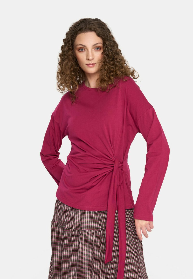 Długi rękaw, głęboki różowy top z węzłem na wysokości talii, wykonany z miękkiego materiału, noszony na spódnicy w kratę z zakładkami.