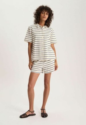 Femme debout portant une chemise crème coordonnée et un short avec de fines rayures horizontales foncées et des sandales noires à bout ouvert sur un fond uni.